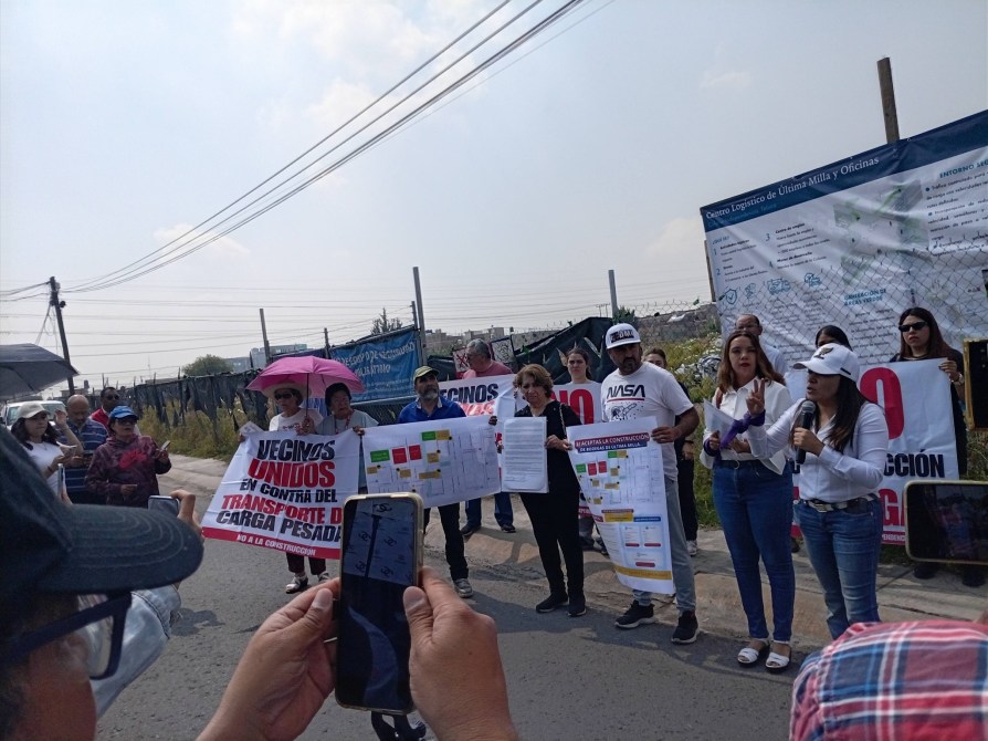 A group of protesters holding signs opposing heavy cargo transport, gathered on a street with a banner in the background. Some participants are using umbrellas for shade.