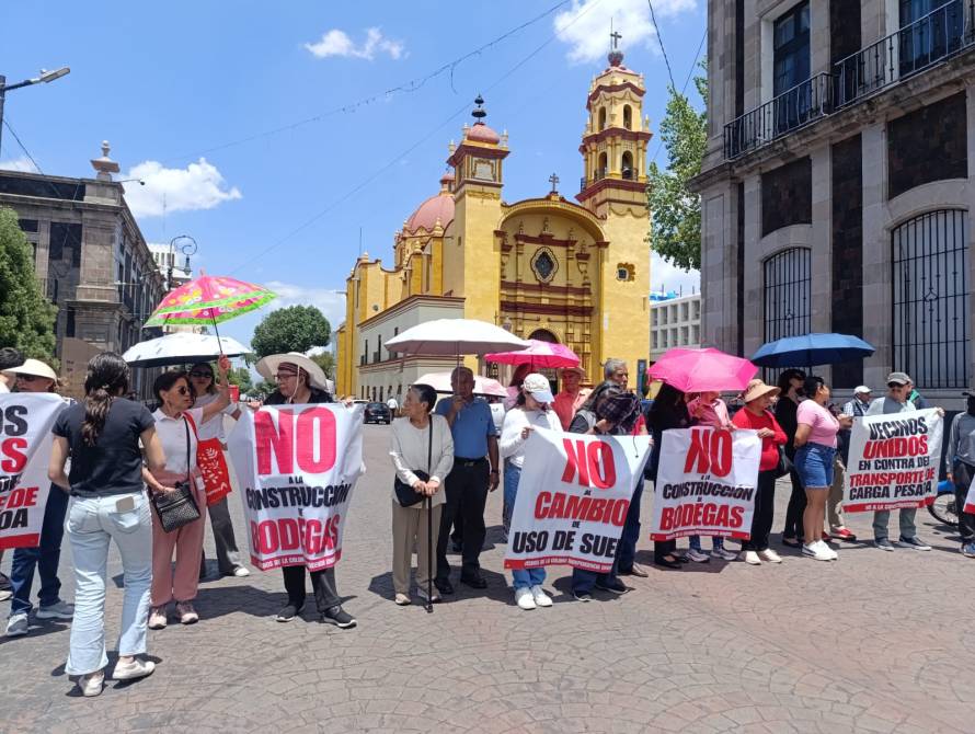 A group of protesters holding signs opposing construction projects, standing in front of a colorful church. Some attendees are using umbrellas for shade on a sunny day.