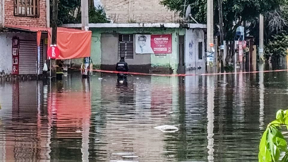 Inundación en una calle con casas parcialmente sumergidas, un hombre sentado en el agua y carteles visibles en las paredes.