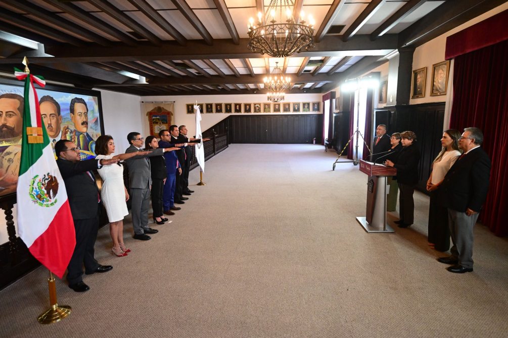 A formal ceremony with officials raising their right hands, taking an oath with the Mexican flag in the foreground. Two women are speaking at a podium, while others are lined up attentively in a large, decorated room.