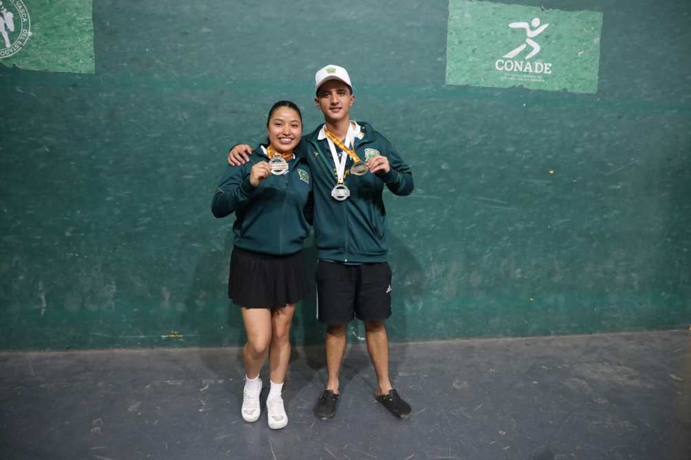 Dos atletas posando juntos, cada uno sosteniendo medallas, en un fondo verde con logotipos deportivos.