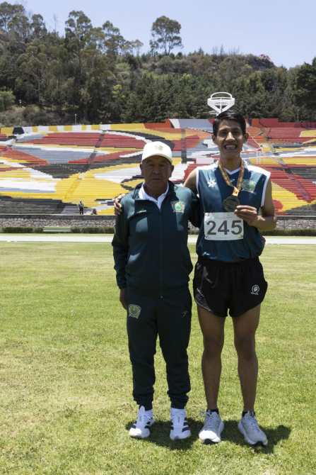 Un atleta sonriente con una medalla de oro junto a su entrenador en un campo deportivo, con gradas coloridas de fondo.