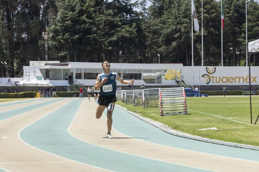 Atleta corriendo en una pista con número 245 en el pecho, celebrando su victoria, con un fondo de instalaciones deportivas y público.