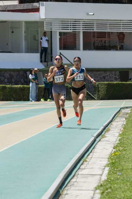 Dos atletas compiten en una pista de atletismo, corriendo lado a lado. La primera atleta lleva el número 372 y la segunda el 213. Se aprecia un ambiente soleado y espectadores al fondo.