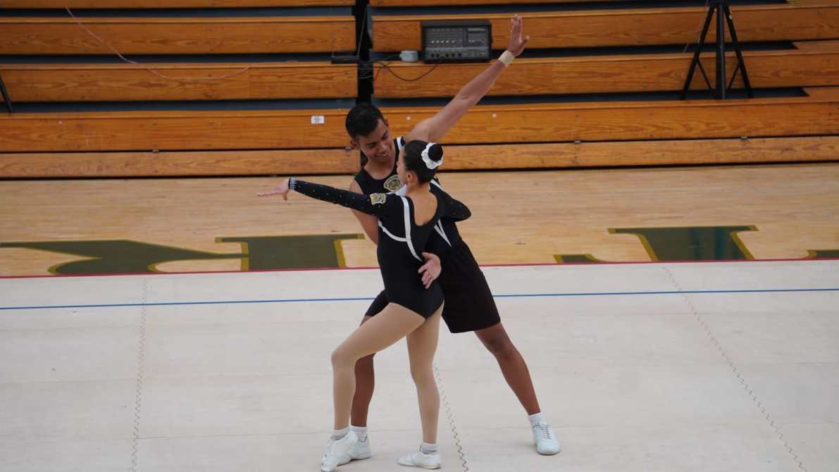 Pareja de bailarines realizando una rutina de baile en una pista de competencia. Ella viste un traje negro con detalles plateados y él lleva una vestimenta similar. Fondo de gradas de madera.