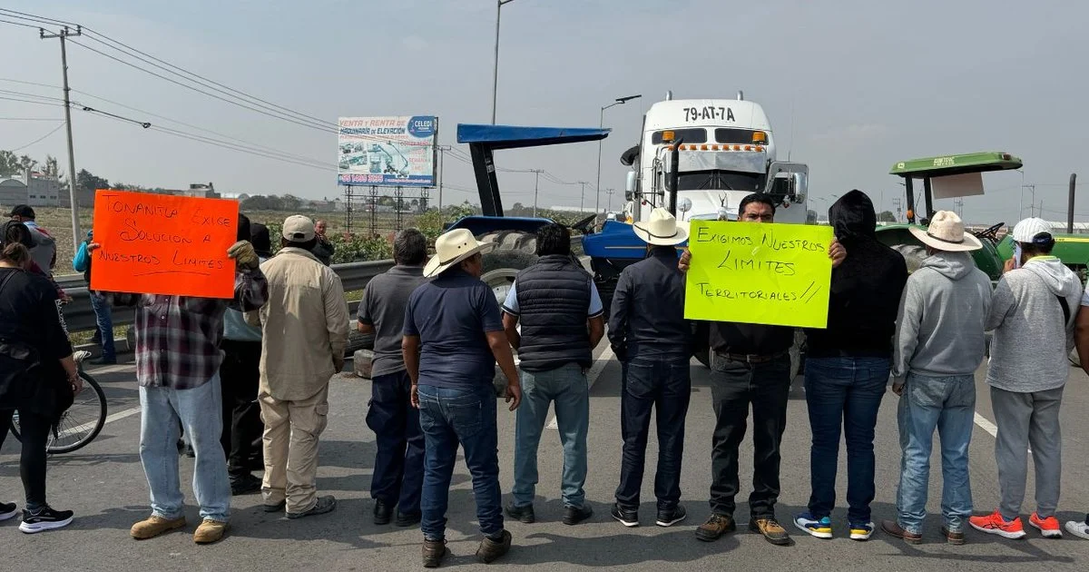 Protesta de personas en la carretera sosteniendo carteles que exigen solución a límites territoriales, con camiones y tractores en el fondo.
