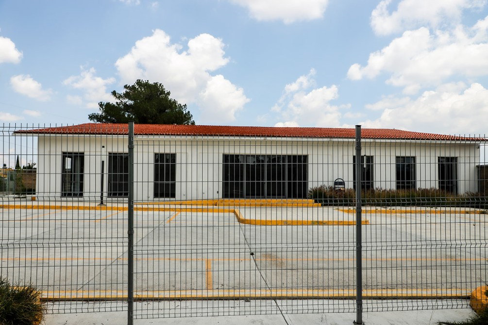 Edificio blanco con techo de tejas, rodeado de una cerca metálica, y un estacionamiento vacío.