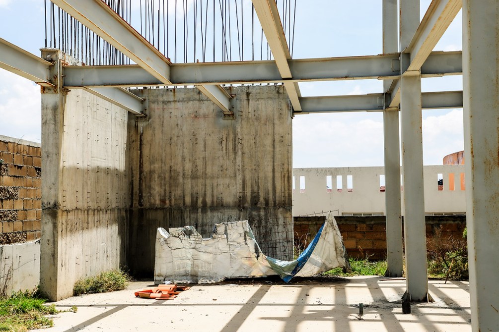 Interior de una construcción en ruinas, mostrando estructuras de acero y paredes de concreto. Hay una lona arrugada en el suelo y escombros alrededor.