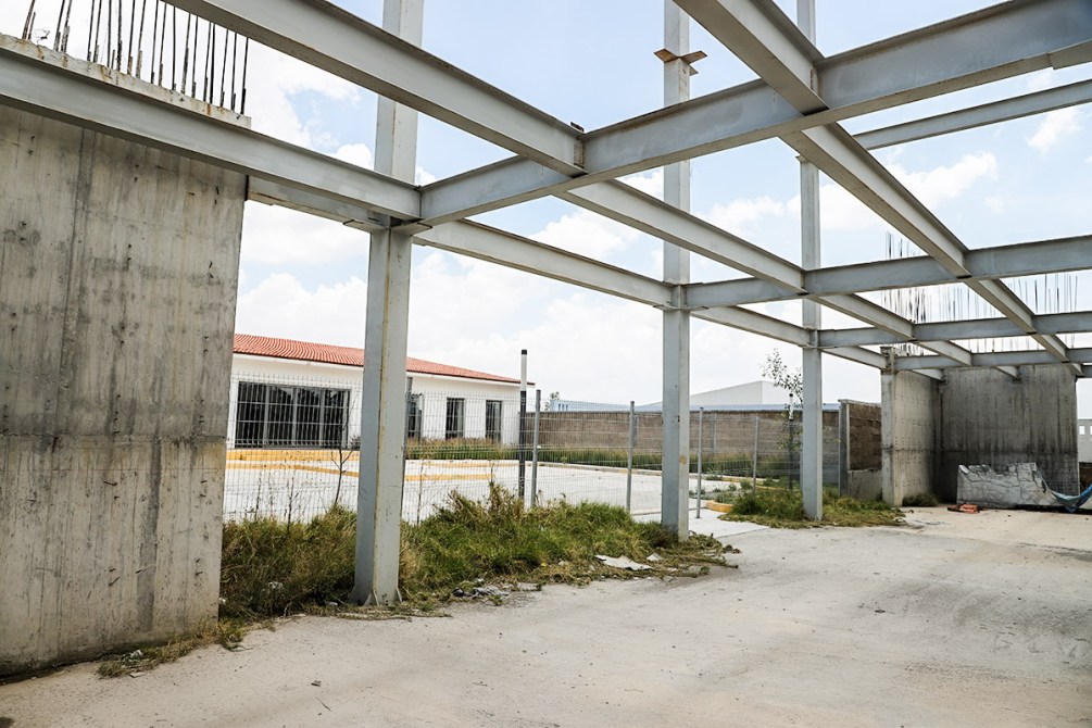 Vista interior de una construcción en proceso, con estructuras metálicas expuestas y paredes de concreto sin terminar, rodeada de hierba y un fondo de edificio y cercas.