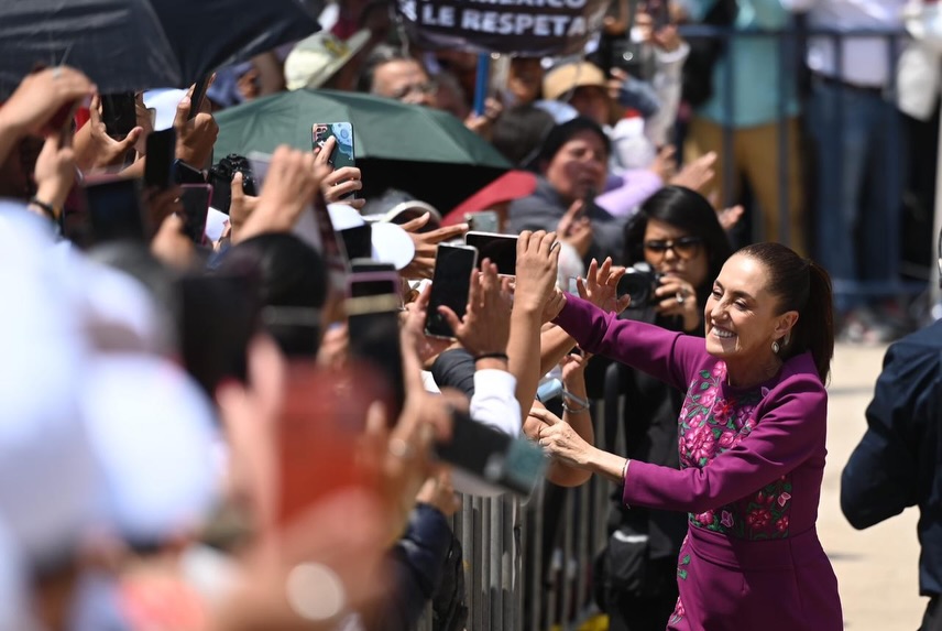 Una mujer sonriente saluda y estrecha manos con una multitud de personas que levantan sus teléfonos en un evento al aire libre.