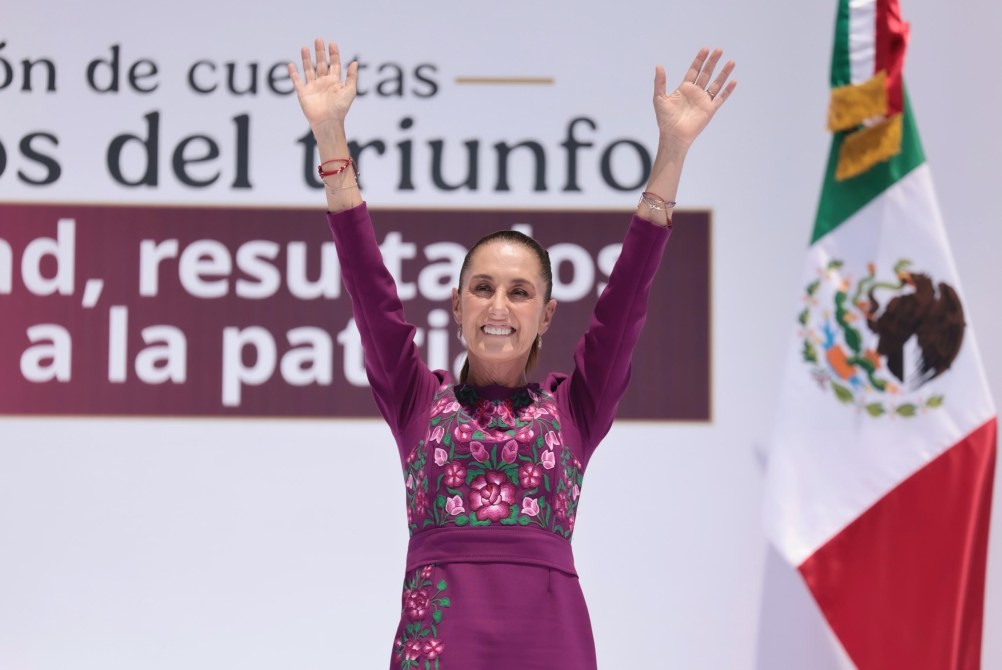Una mujer sonriente levantando ambas manos en señal de triunfo, vestida con un atuendo tradicional mexicano, se encuentra frente a un fondo con un cartel que dice 'Cuentas del triunfo'. A la derecha, se puede ver la bandera de México.
