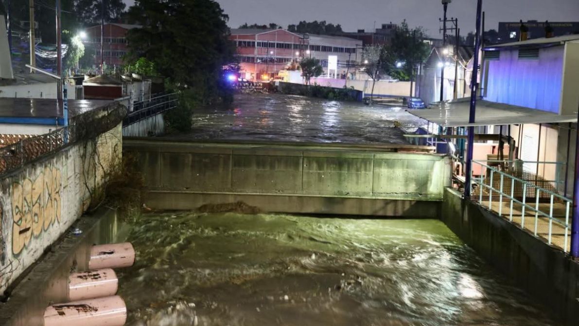 Inundación nocturna en una zona urbana con un río muy crecido, con signos de desbordamiento. Se observan edificios y luces de emergencia en el fondo.