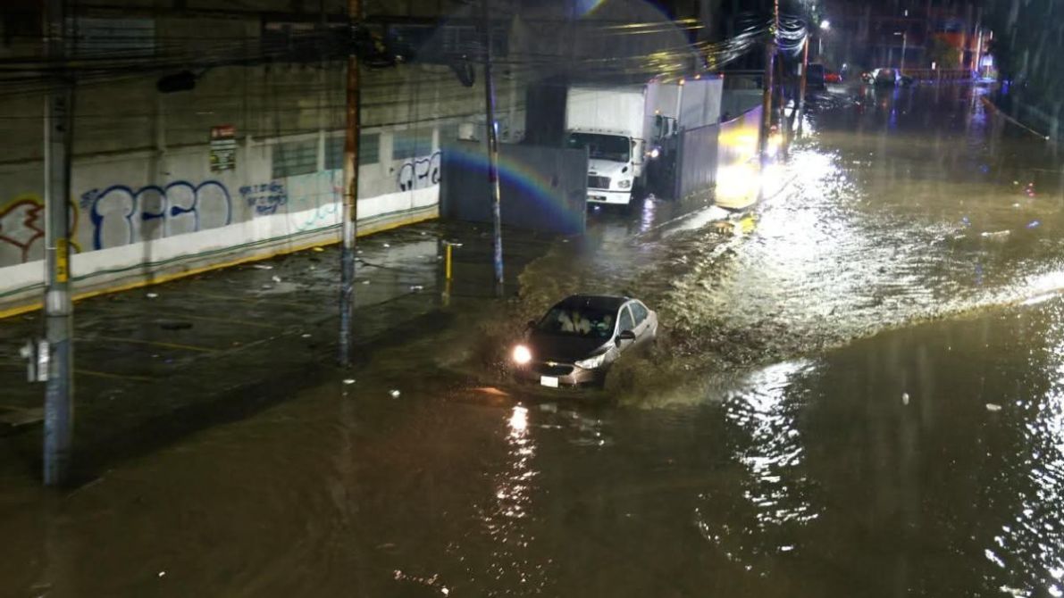 Un automóvil atravesando una calle inundada por fuertes lluvias, con agua que cubre gran parte de la vía y vehículos estacionados parcialmente sumergidos.
