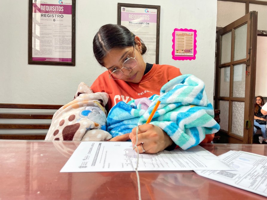 Una joven sentada en un escritorio, escribiendo en un formulario mientras sostiene mantas de colores en sus brazos.