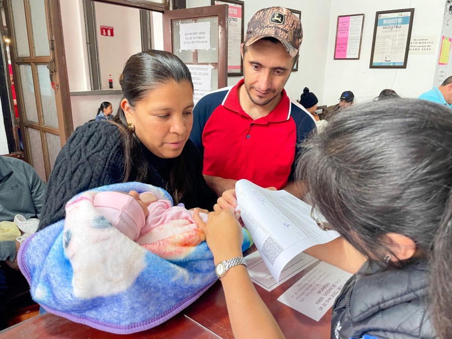 Una familia en una oficina, la madre sostiene a un bebé en una manta mientras discuten con un funcionario que revisa documentos.