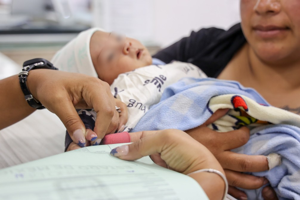 Una madre sostiene a su bebé en brazos mientras un profesional de la salud toma notas y mide al infante en un entorno médico.