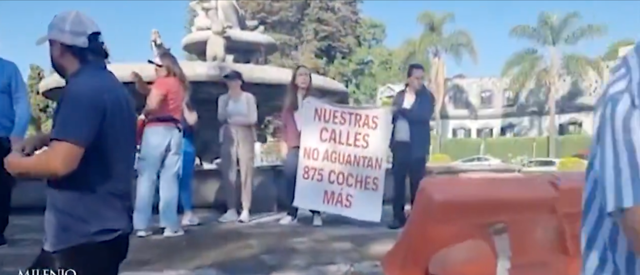 Manifestantes sosteniendo un cartel que dice 'Nuestras calles no aguantan 875 coches más', frente a una fuente en un parque.