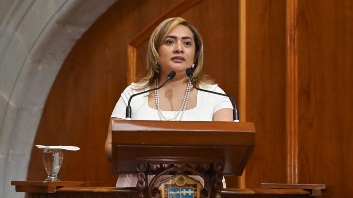 Mujer haciendo un discurso en un podio en un entorno institucional con paneles de madera de fondo.