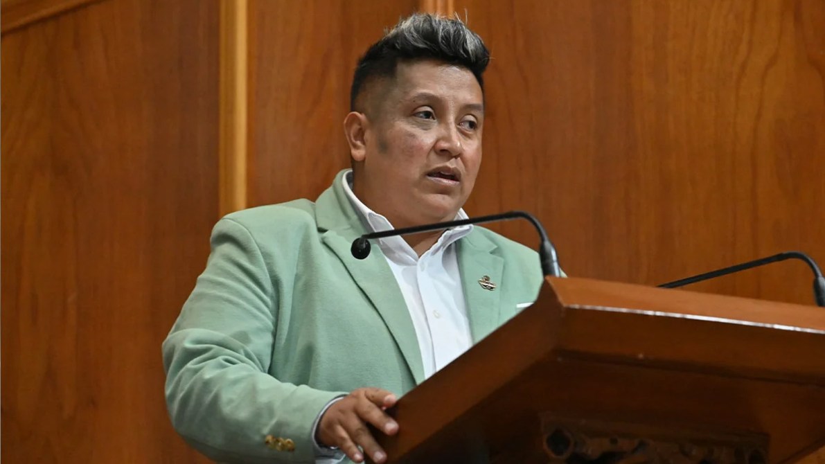 Una persona hablando en un podio, vestido con un saco verde claro y una camisa blanca, frente a un fondo de madera.