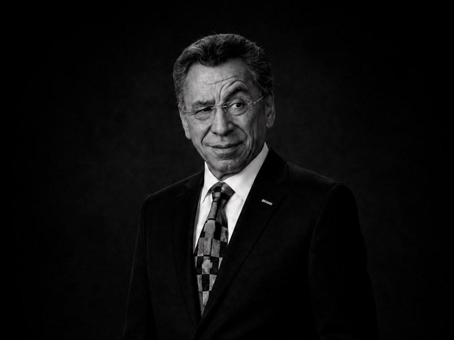 Retrato en blanco y negro de un hombre mayor con gafas, vestido con un traje negro y una corbata de colores, sonriendo ligeramente frente a un fondo oscuro.
