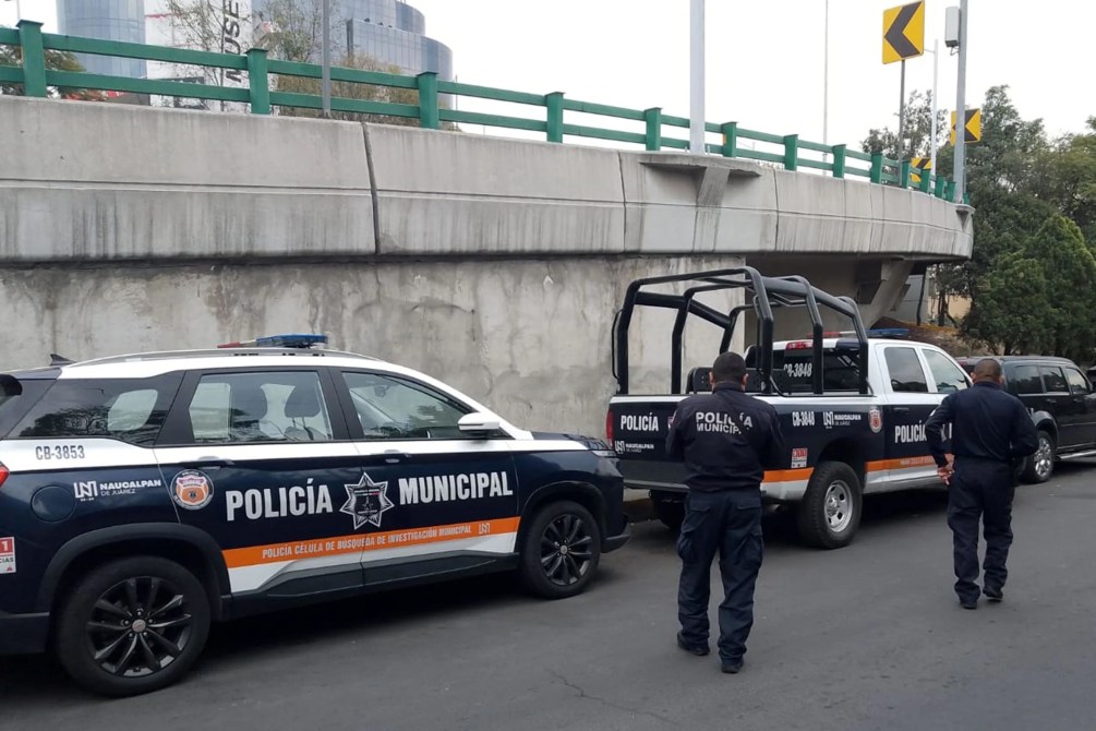 Two municipal police officers standing next to police vehicles parked on the side of the road, with a concrete overpass in the background.