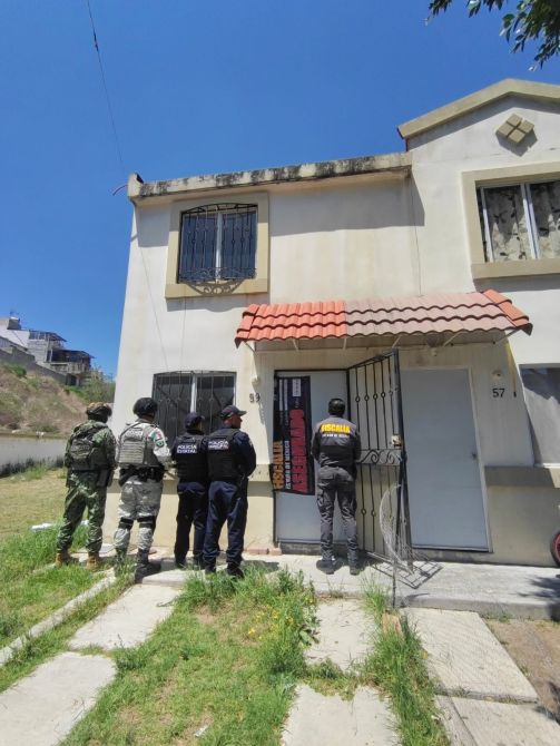 Agentes de seguridad y un militar frente a una casa, observando la puerta principal.