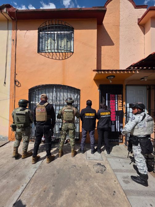 Grupo de agentes de seguridad frente a una casa con puertas cerradas, algunos vestidos con uniformes tácticos y otros con prendas que llevan el sello de la fiscalía.