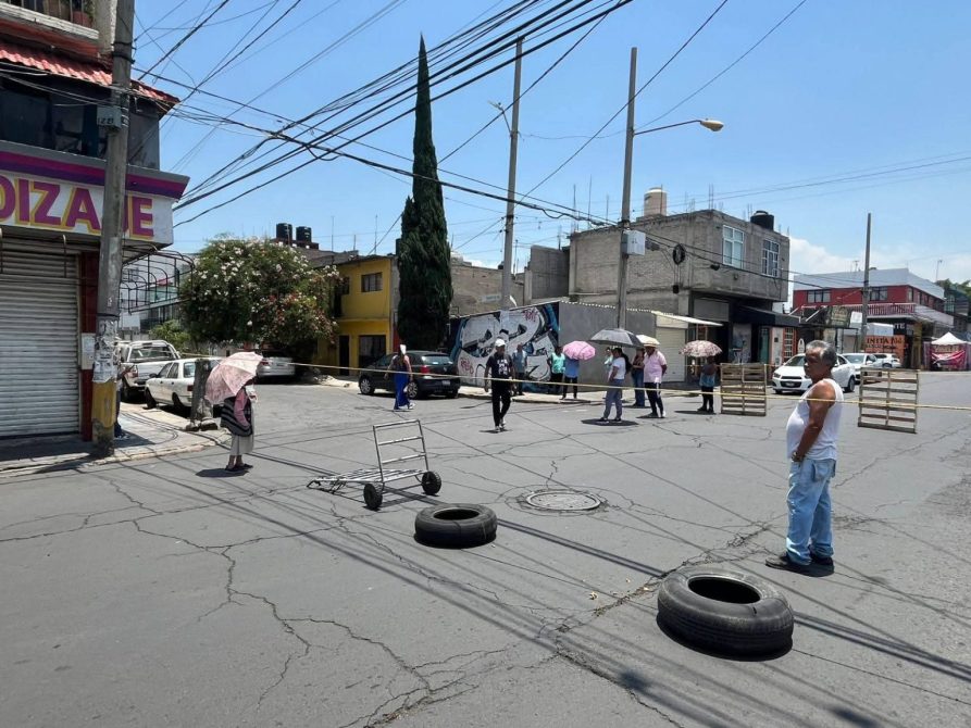 Una calle con personas caminando bajo paraguas, un carro de compras vacío y neumáticos en el suelo, rodeado de edificios y vegetación.