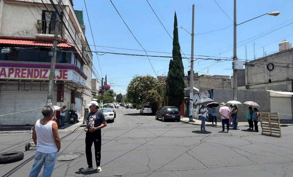 Escena de una calle en una zona urbana con edificios y árboles. Varias personas están de pie, algunas usando sombrillas, mientras que un par de hombres conversan en el cruce. Se observan vehículos estacionados y un cielo despejado.