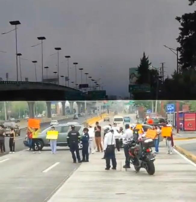 Protesta en una carretera con varias personas sosteniendo pancartas y vehículos detenidos. Se ven policías presentes y un ambiente nublado.