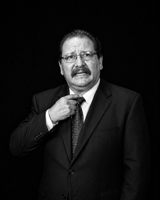 Un hombre de mediana edad con bigote, usando un traje negro y una corbata, parece preocupado mientras ajusta su corbata sobre un fondo negro.