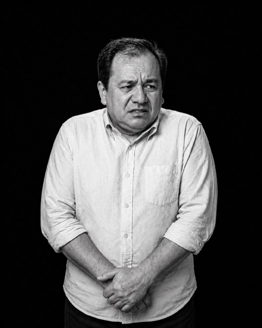 Hombre con expresión de incomodidad, de pie frente a un fondo negro, con las manos cruzadas y mirada seria.
