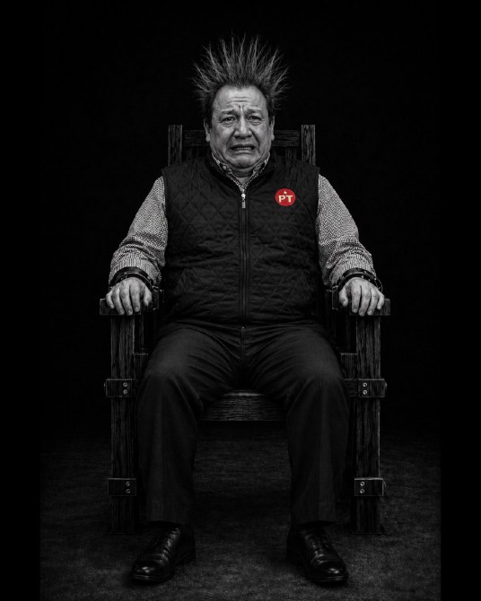 Retrato en blanco y negro de un hombre sentado en una silla, con cabello erguido y expresión facial seria, vestido con un chaleco negro con el logo del PT.