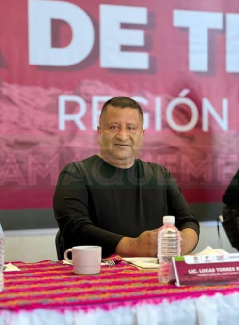 Hombre sentado en una mesa con una camisa negra, sonriente, en un evento con un fondo que dice 'Tierra de Tequila Región'.