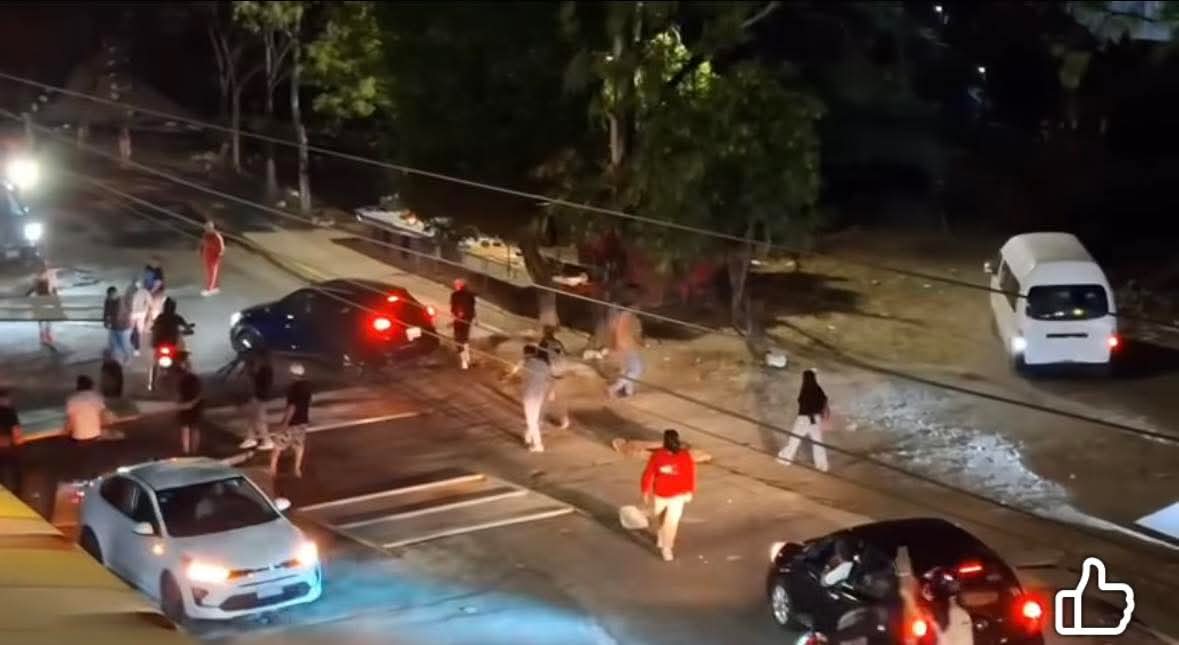 Grupo de personas en una calle durante la noche, algunas caminando y otras en vehículos, con luces de coches visibles.