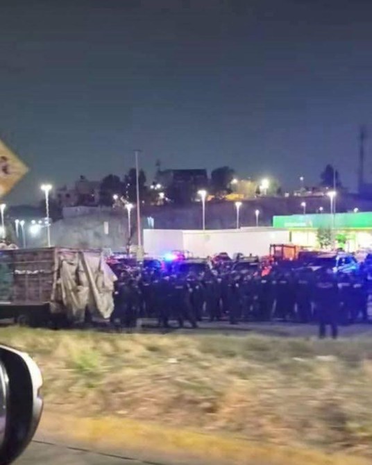 Una gran cantidad de policías en una carretera, con luces azules y rojas de vehículos patrullando, durante la noche.