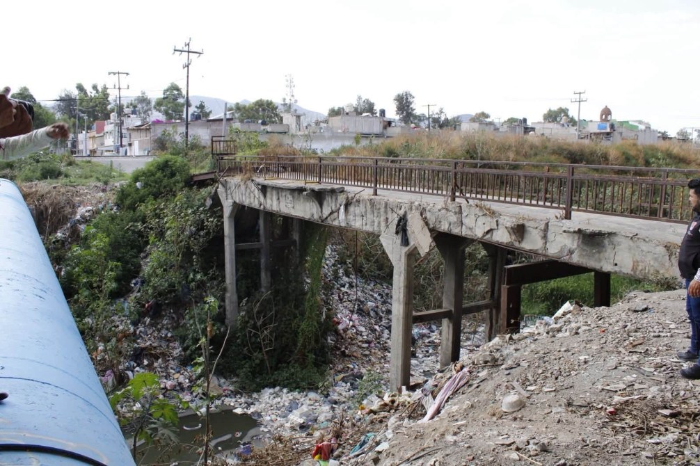 Puente en mal estado sobre un arroyo contaminado, rodeado de basura.