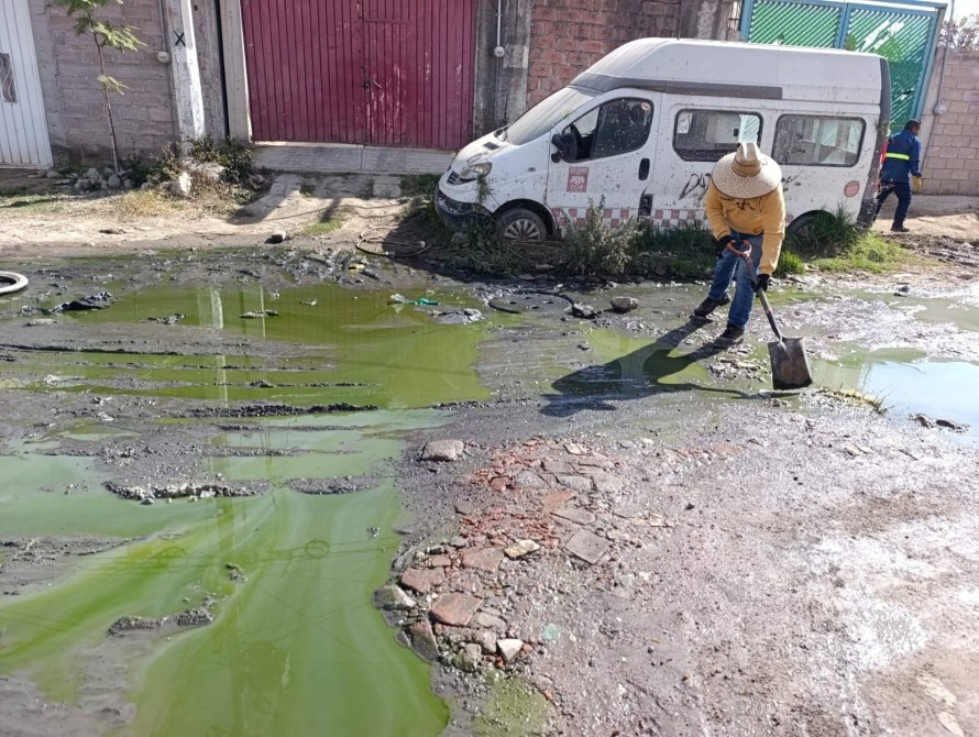 Hombre trabajando en una calle con suelo en mal estado y charcos de agua verde, junto a una camioneta abandonada.