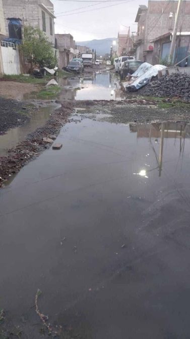 Vista de una calle inundada con agua estancada, vehículos estacionados y casas a los lados.