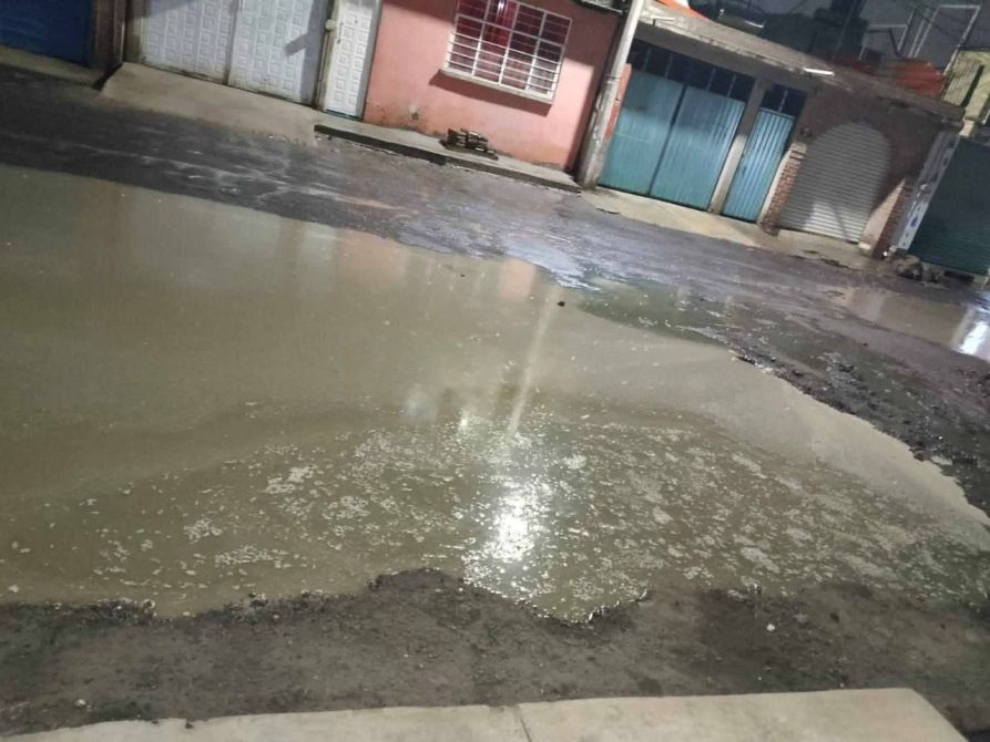 Charco de agua sucia en una calle con casas y garajes de fondo.