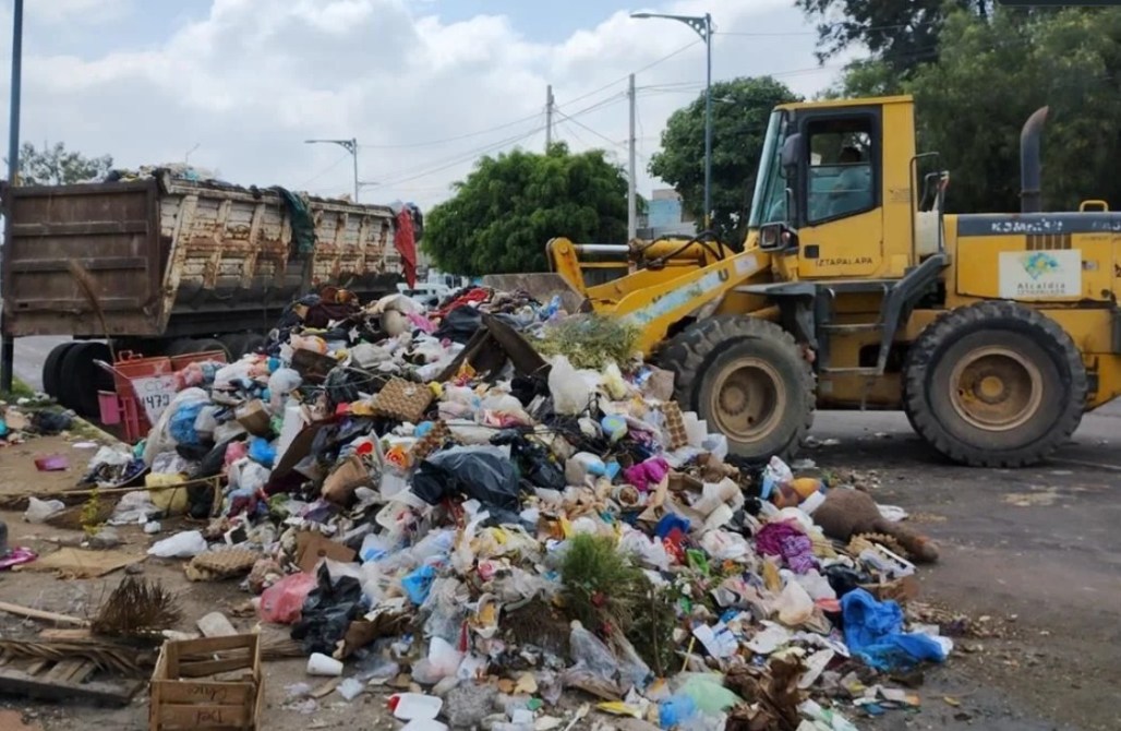 Montón de basura junto a un camión de carga y una retroexcavadora en una calle.