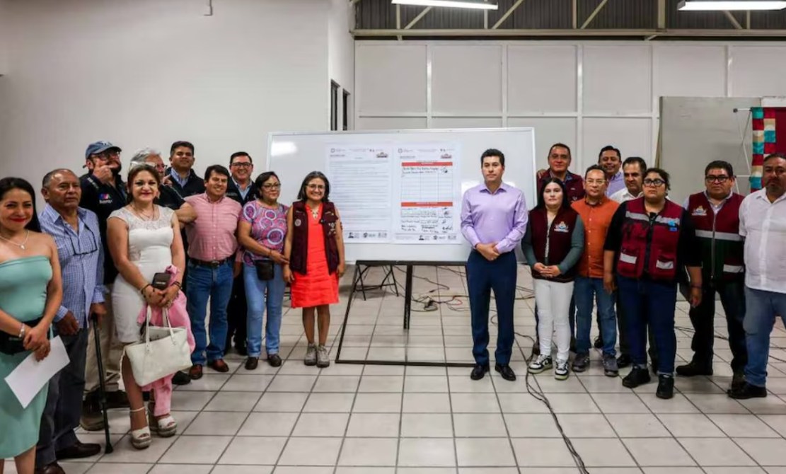 Grupo de personas posando frente a una presentación en un salón. Hay un pizarrón con documentos en el fondo y todos los participantes sonríen, mostrando un ambiente colaborativo.