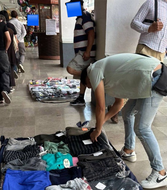 Una persona se agacha para revisar ropa en el suelo mientras otras personas están de pie en un pasillo con mercancías.