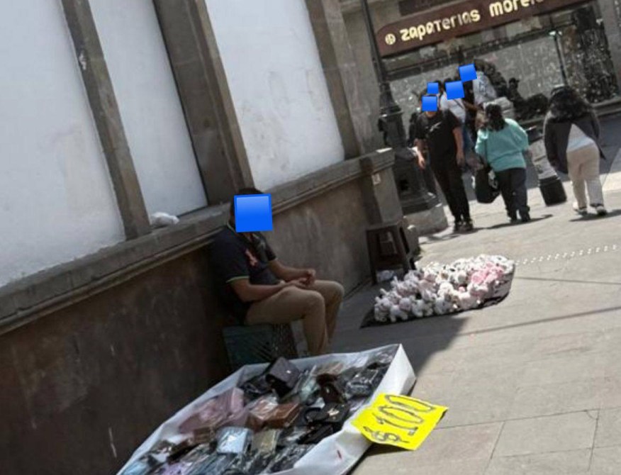 Una persona sentada en la acera al lado de una manta con artículos en venta, mientras otras personas caminan por la calle.