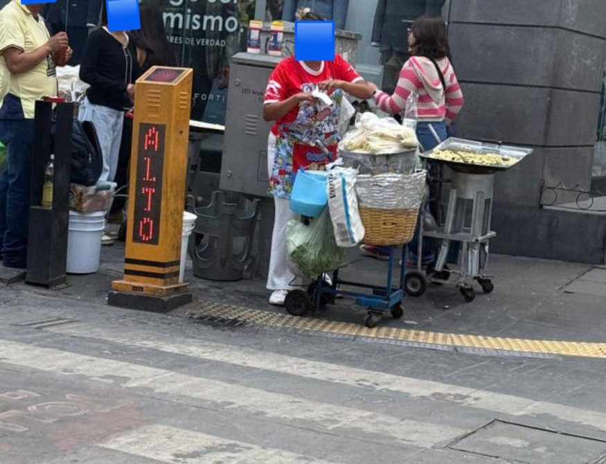 Una vendedora ambulante en la calle preparando comida, con un carrito lleno de ingredientes y una señal LED que dice 'ALTO'.