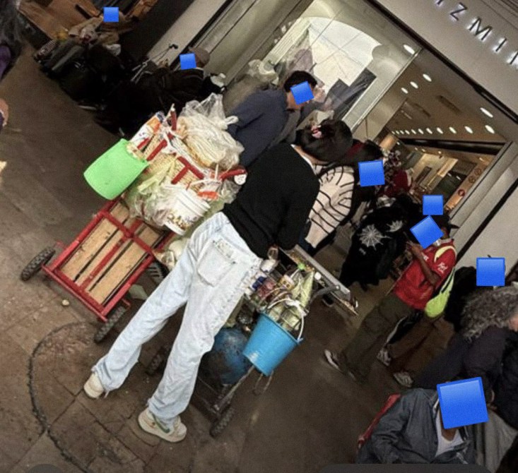 Persona vendiendo comida en un mercado, con un carrito lleno de productos y varios clientes en las cercanías.