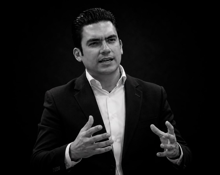 Hombre de pie en un entorno oscuro, gesticulando con las manos mientras habla con pasión, vestido con un traje oscuro y una camisa blanca.