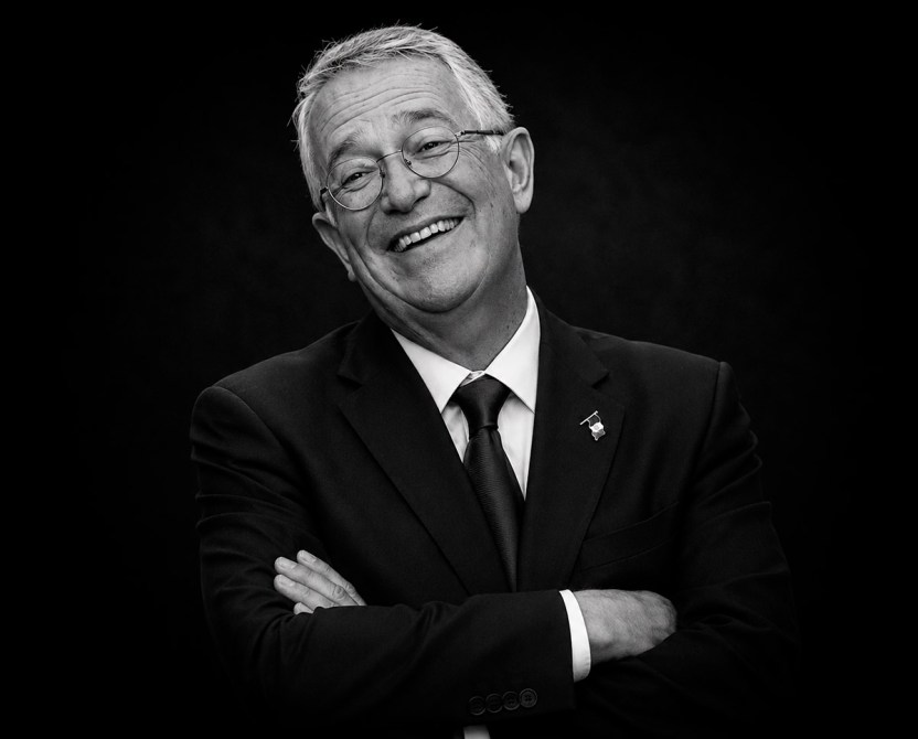 Retrato en blanco y negro de un hombre con gafas y una sonrisa, vestido con un traje negro y corbata, posando con los brazos cruzados.