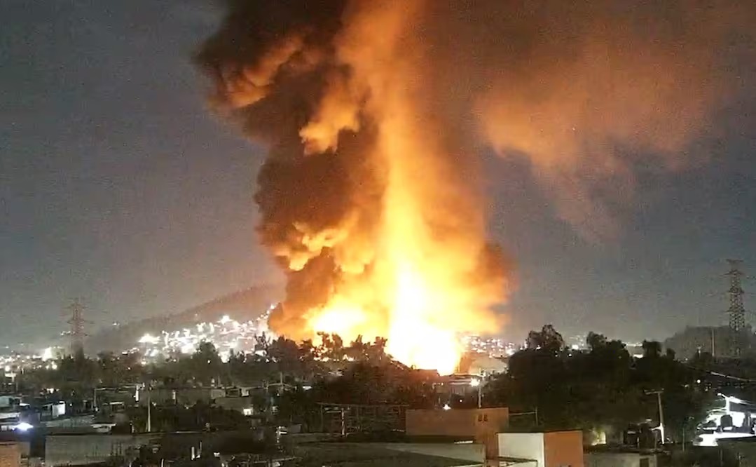 Incendio masivo durante la noche con una gran columna de humo y llamas visibles, afectando una zona urbana.