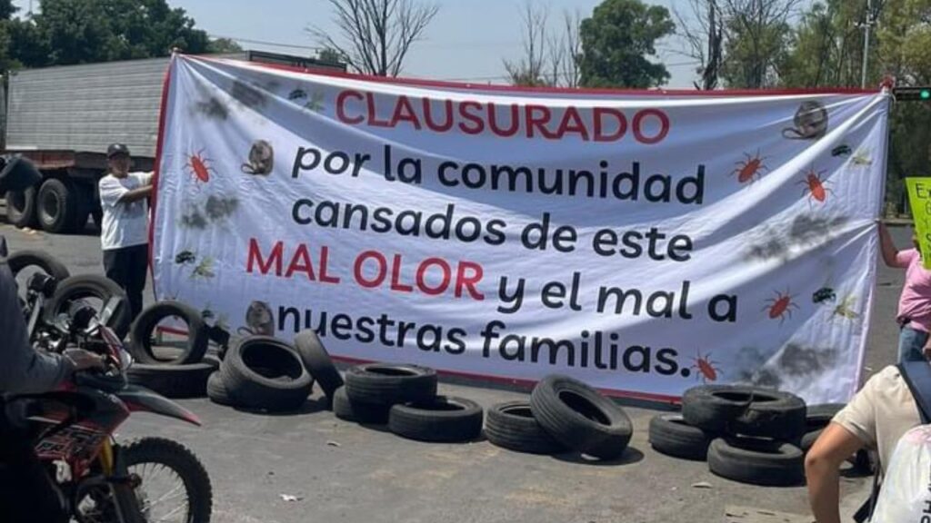 Protesta comunitaria con un gran cartel que dice 'CLASURADO por la comunidad, cansados de este MAL OLOR y el mal a nuestras familias', junto a neumáticos en el suelo.